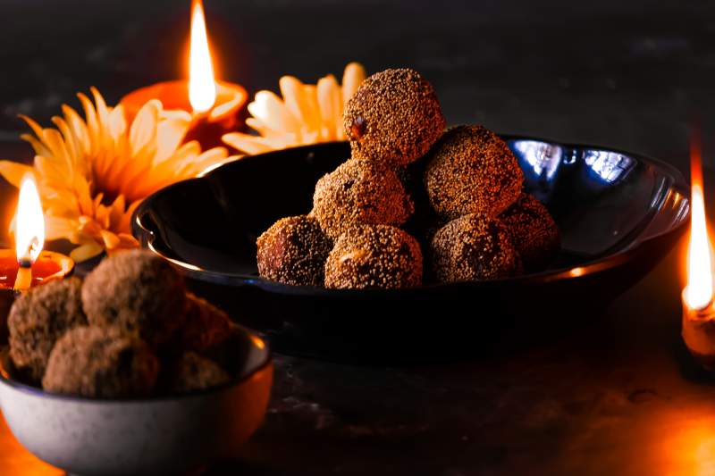 A black bowl with ladoo and another white bowl with ladoo and diyas and flowers in the background