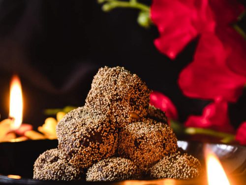 a bowl with ladoo and diya in the front and back with red flower in the background