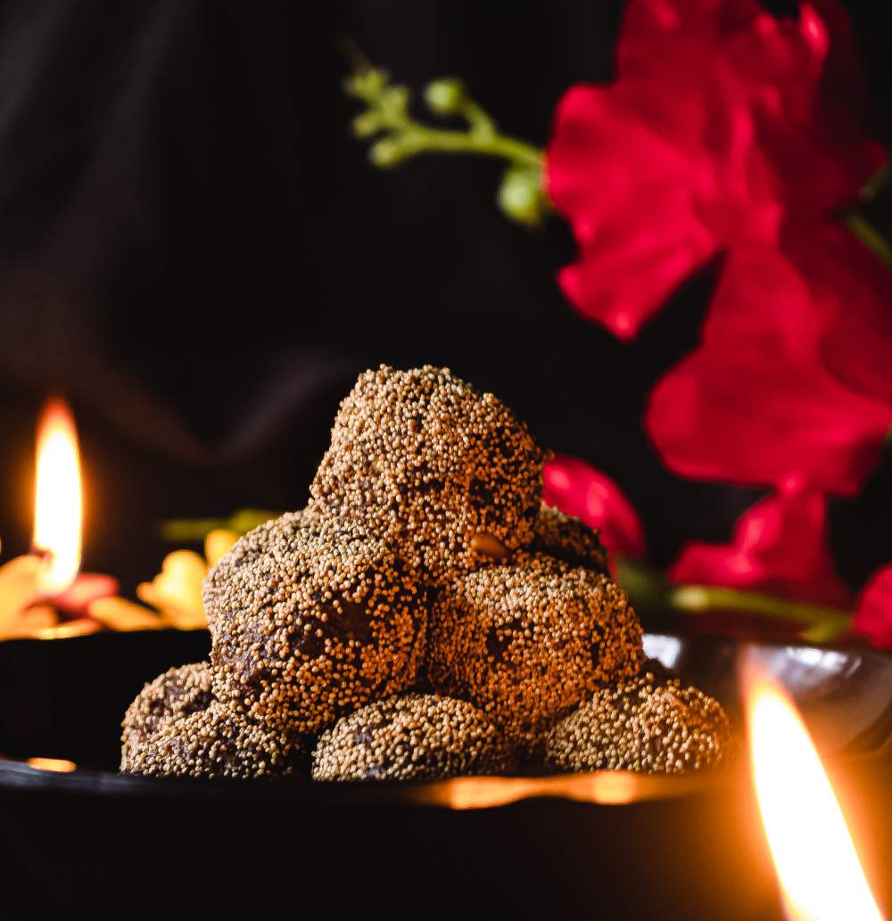 a bowl with ladoo and diya in the front and back with red flower in the background